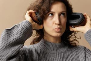 Pensive funny curly beautiful female in gray casual sweater headphones listen to her recording in studio posing isolated on over beige pastel background. Sound streaming platform concept. Copy space photo