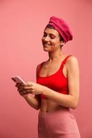 joven mujer blogger con de colores rosado pelo y corto Corte de pelo voltear mediante el pantalla de el teléfono y mecanografía un mensaje con un sonrisa en elegante ropa en un rosado antecedentes foto