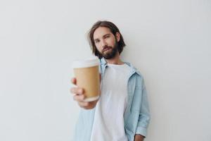 Lanza libre milenario hombre con un barba Bebiendo café desde un reciclado taza en elegante hipster ropa blanco camiseta azul pantalones y camisa en un blanco antecedentes foto