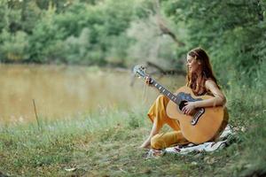 hippie mujer jugando guitarra sonriente y canto canciones en naturaleza sentado en un tartán por el lago en el noche en el rayos de el ajuste Dom. un estilo de vida en armonía con el cuerpo y naturaleza foto