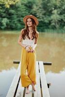 A young woman in a hippie look and eco clothing travels outdoors by the lake wearing a hat and yellow pants in the fall photo