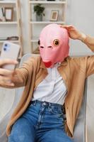 Happy funny blonde lady in pink fish mask warm sweater doing selfie video call sitting in armchair at modern home interior. Pause from work, take a break, social media in free time concept. Copy space photo