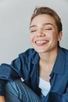 un joven mujer sentado en un silla a hogar sonriente con dientes con un corto Corte de pelo en pantalones y un mezclilla camisa en un blanco antecedentes. niña natural poses con No filtros foto