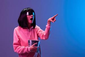 Excited awesome brunet woman in pink hoodie trendy specular sunglasses with popcorn point finger aside posing isolated in blue violet color light background. Neon party Cinema concept. Copy space photo