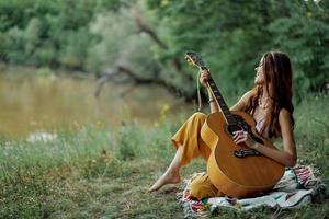 un hippie mujer jugando su guitarra sonrisas y canta canciones en naturaleza sentado en un tartán en el noche en el puesta de sol luz de sol. un estilo de vida en armonía con el cuerpo y naturaleza foto
