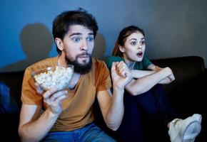un hombre y un asustado mujer acecho un película en el noche en el sofá con un plato de palomitas de maíz foto