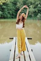 A young woman in a hippie look and eco-dress dancing outdoors by the lake wearing a hat and yellow pants in the summer sunset photo