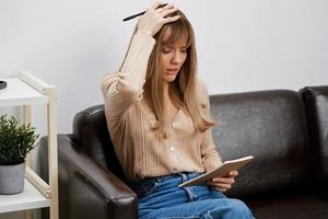 Upset confused unhappy blonde student lady with copybook touch head sitting in sofa couch at modern living room interior. Female client have shrink appointment in psychological help office. Copy space photo