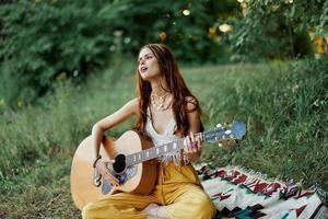 joven hembra hippie artista obras de teatro guitarra y canta canciones en Respetuoso del medio ambiente ropa sentado en el suelo fuera de en naturaleza en el otoño mirando fuera a el puesta de sol foto