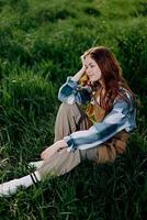 A young, beautiful woman lounges on the green grass in the park wearing sneaker pants and a plaid shirt and looks out into the setting sun photo