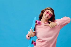 Cute smiling pretty redhead lady in pink hoodie sunglasses with penny board look at camera posing isolated on blue studio background. Copy space Banner Offer. Fashion Cinema. Holiday activity photo