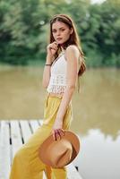 A young woman smiling in an image of a hippie and eco-dress dancing in nature by the lake wearing a hat and yellow pants in the summer sunset photo