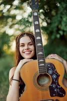 joven hippie mujer con eco imagen sonriente y mirando dentro el cámara con guitarra en mano en naturaleza en un viaje foto