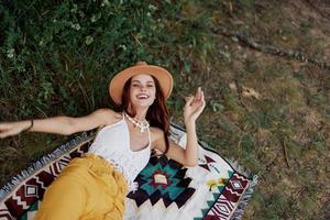 un mujer en Respetuoso del medio ambiente ropa en un hippie Mira es acostado en un de colores tartán sonriente y mirando a un otoño puesta de sol en naturaleza en el parque. estilo de vida en un viaje de armonía con el mundo foto