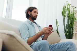 A freelance man in a white T-shirt, blue jeans, and shirt sits on the couch with his phone at home on his day off and plays games photo