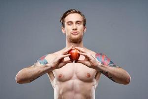 a guy with a pumped-up torso and a tattoo holds an apple in his hands on a gray background photo