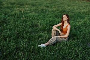 The redheaded woman sits in the park on the green grass wearing an orange top, green pants, and sneakers and looks out at the setting summer sun photo