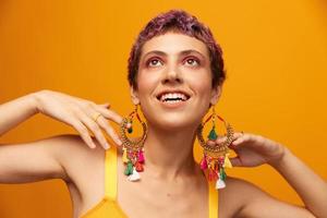 retrato de un joven mujer con un corto Corte de pelo y de colores pelo sonriente y demostración su lengua a el cámara en un naranja antecedentes con pendientes accesorios en el estudio foto