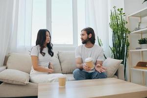 Man and woman sitting at home on the couch in white stylish t-shirts drinking coffee out of crab cups from a coffee shop and having fun chatting smiles and laughter at home. Male and female friendship photo