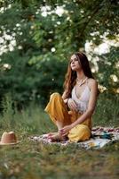 A young hippie woman meditates in nature in the park, sitting in a lotus position on her colorful plaid and enjoying harmony with the world in eco-clothing in the autumn photo