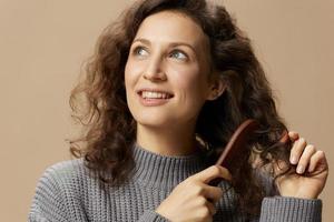 Cheerful curly beautiful female in gray casual sweater with hairbrush combs hair looks aside posing isolated on over beige pastel background. Problematic unruly damaged hair concept. Copy space photo