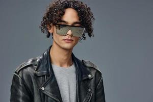 Stylish tanned curly man leather jacket trendy mirror sunglasses posing isolated on over gray studio background. Cool fashion offer. Huge Seasonal Sale New Collection concept. Copy space for ad photo