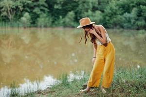 A young woman in a hippie look travels in nature by the lake wearing a hat and yellow pants in the fall photo