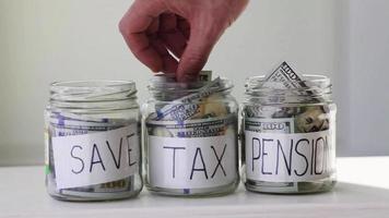 Male hand puts 100 dollars in a glass jar with the text  tax , save, pension . Concept of saving money. video