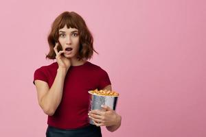 Excited shocked cute redhead lady in red t-shirt with popcorn ready for movie evening posing isolated on over pink studio background. Copy space Banner. Fashion Cinema concept. Entertainment offer photo