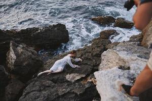 Top-down aerial view of beautiful young woman in long white dress lying on rocky coast with cracks on rocky surface photo