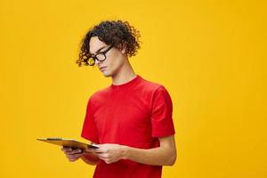 Thinking myopic young student man in red t-shirt funny eyewear holds tablet folder with study notes posing isolated on over yellow studio background. Free place for ad. Education College concept photo