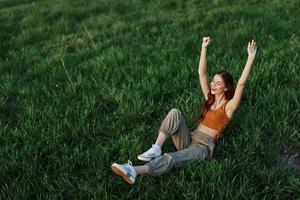 un joven mujer es jugando en el parque con nosotros en el césped y caídas a el suelo sonriente felizmente en el luz de sol. descansando en naturaleza, armonía con el cuerpo foto