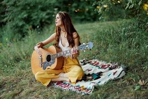 joven hembra hippie artista obras de teatro guitarra y canta canciones en Respetuoso del medio ambiente ropa sentado en el suelo fuera de en naturaleza en el otoño mirando fuera a el puesta de sol foto