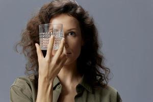 Pretty curly beautiful woman in casual khaki shirt hold glass of water near face close an eye posing isolated on over gray blue background. Healthy lifestyle. Water is Life concept. Abstract shot photo