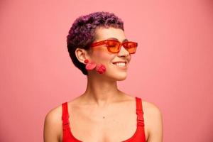 Moda retrato de un mujer con un corto Corte de pelo en de colores Gafas de sol con raro accesorios con pendientes sonriente en un rosado brillante antecedentes foto