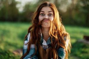 gracioso mujer retrato de cerca riendo y meneando para el cámara en frente de un verde parque en verano, el puesta de sol brillante en su rojo pelo foto