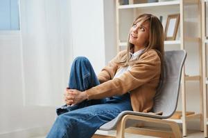 Cheerful dreaming young blonde lady in warm sweater looks up aside sitting in armchair at modern home interior. Pause from work, take a break, reflections in free time concept. Copy space photo