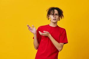 Confused funny Caucasian young man in red t-shirt point both hands aside posing isolated on over yellow studio background. The best offer with free place for advertising. Emotions for everyday concept photo
