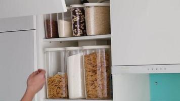 A male hand opens and closes a cabinet in a modern white kitchen without handles. Stocked kitchen pantry with food - pasta, buckwheat, rice and sugar . The organization and storage in kitchen. video
