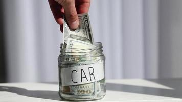 Male hand puts 100 dollars in a glass jar with the text car. Concept of saving money. video