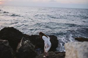 barefoot woman in a white dress with wet hair stands on a cliff photo