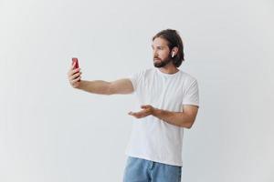 A man with a beard blogger in a white T-shirt with a phone and wireless headphones talking on an online video call against a white background photo