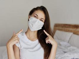 happy woman sitting on bed and showing finger to medical mask on face photo