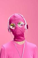 A crazy conceptual portrait of a young woman without a face mask on a pink background and wearing shiny sunglasses listening to music on her headphones. The concept of a new world and cyberspace photo