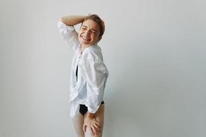 joven mujer adolescente escuchando a música con estéril auriculares y bailando hogar, sonriendo con dientes con un corto Corte de pelo en un blanco camisa en un blanco antecedentes. niña natural poses con No filtros foto