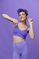 Deportes Moda mujer bailando posando sonriente con dientes en un púrpura Deportes traje para yoga en un esbelto cuerpo y un transparente gorra en su cabeza en un púrpura antecedentes monocromo foto