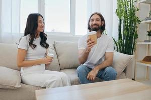hombre y mujer sentado a hogar en el sofá en blanco elegante camisetas Bebiendo café fuera de cangrejo tazas desde un café tienda y teniendo divertido chateando sonrisas y la risa a hogar. masculino y hembra amistad foto