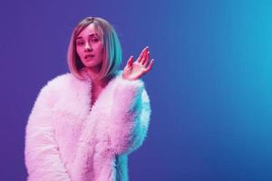 Confused excited adorable blonde woman in fluffy fur coat sparkly dress show hand up posing isolated in blue violet pink color light studio background. Neon party Fashion concept. Copy space Banner photo