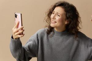 Crying scandalising curly beautiful female in gray casual sweater do video call with husband posing isolated on over beige background. Social media, network, distance communication concept. Copy space photo