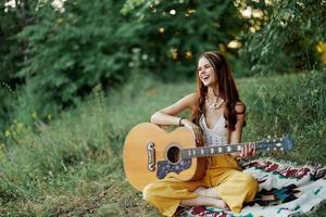 joven hembra hippie artista obras de teatro guitarra y canta canciones en Respetuoso del medio ambiente ropa sentado en el suelo fuera de en naturaleza en el otoño mirando fuera a el puesta de sol foto